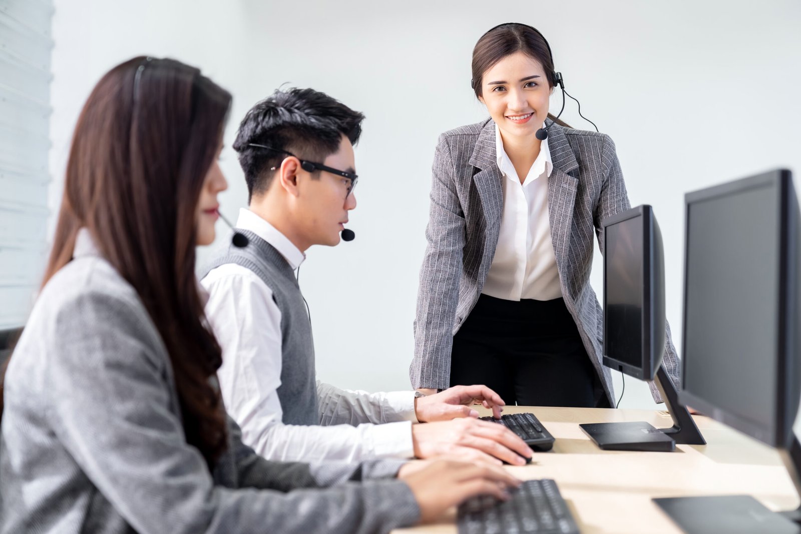 Young adult confidence with smiling female supervisor standing in front of working table of operator team agent with headsets and desktop computer in a call center to monitor her employee. Using for woman leadership concept