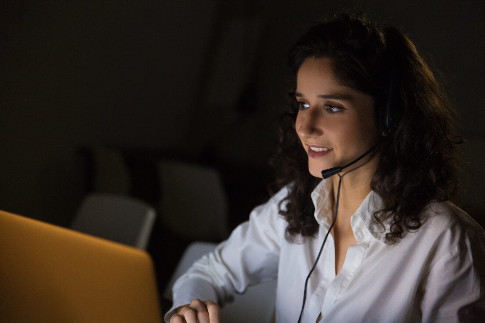 Smiling woman with headset in dark office. Cheerful female call center worker working with laptop late at night. Working late concept